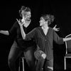 Programmes image for "Character Connection Through Improv". Two woman look at each other. They sit on the same chair. Their hands are thrown up as if shrugging. 