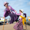Poster image for "The Grannies". Two actors dressed in purple old woman clothes. They each have a cane. One actor is standing acrobatically on the other's knees.